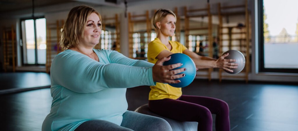 Older lady exercises with personal trainer in a gym with yoga balls as part of her holistic medical weight loss care plan