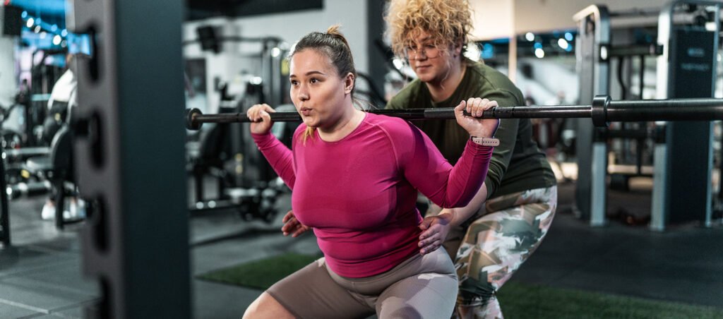 Two women working out together in a gym doing heavy squats to lose weight, build muscle, and improve health along their GLP-1 journey with prescription Ozempic (Semaglutide)