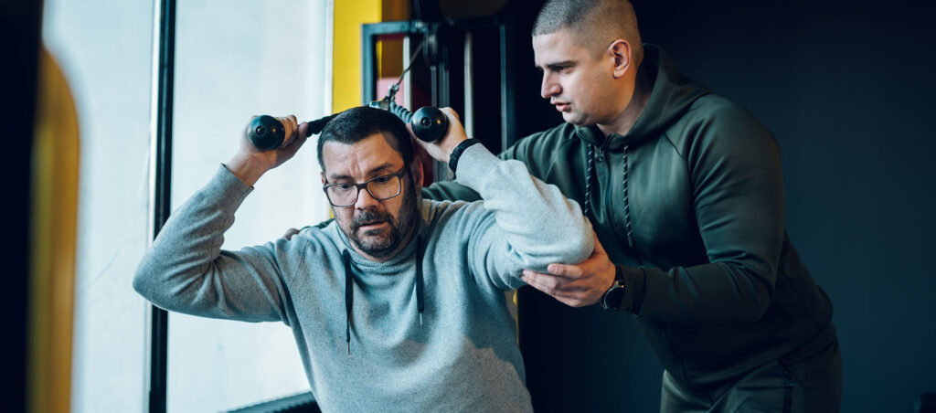 Man taking GLP-1 medication for weight loss works with personal trainer in a gym setting to perfect his strength training technique