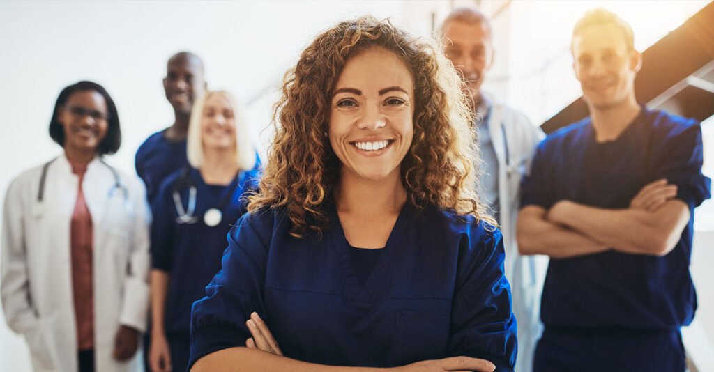 A smiling female nurse stands with her arms crossed in front of her diverse medical team, illustrating a positive approach to sustainable hospital employee wellness.