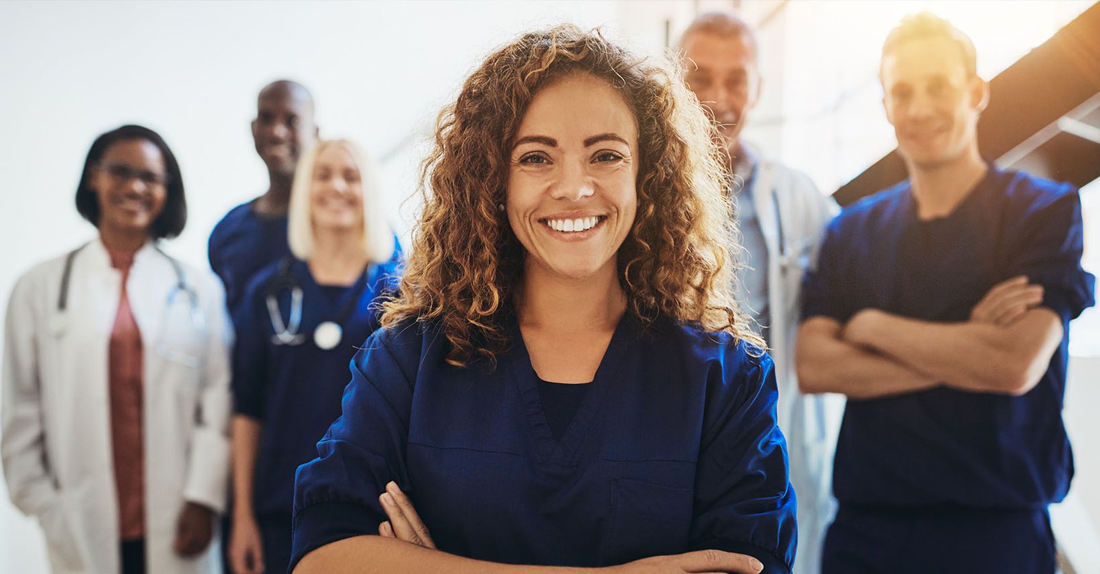 A smiling female nurse stands with her arms crossed in front of her diverse medical team, illustrating a positive approach to sustainable hospital employee wellness.